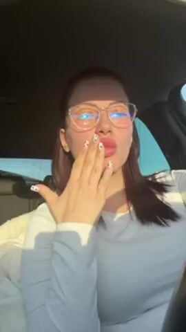 A woman wearing glasses is in the driver's seat of a car. She is wearing a white shirt. The sunlight is shining into the car. She blows a kiss at the camera. She then rests her head on her hand and looks toward the camera.