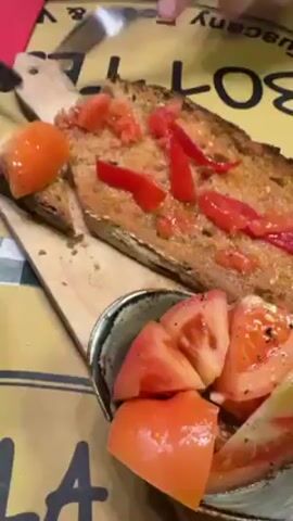 The video shows a wooden platter with a slice of toasted bread topped with red pepper strips and diced tomatoes. Beside it is a small bowl containing tomato slices seasoned with salt and pepper. A person's hands are visible, holding a fork and moving one of the tomato slices from the bowl to the bread. The setting appears to be a restaurant, with a table cover partially visible with text “Food & V” and “BOT” on it. The room is slightly noisy.