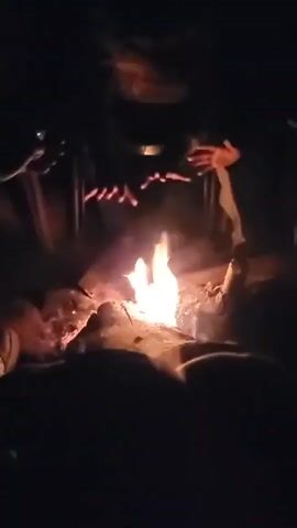 The video captures a dark, nighttime scene of several people gathered around a campfire in Khir Ganga, India. The bright flames of the fire are the primary source of illumination, casting a warm glow on the immediate surroundings and the hands of those present.

The main activity involves individuals holding long, thin food items, possibly sausages or similar snacks, directly over the open flames to roast them. Multiple hands are visible, carefully positioning and turning these items above the fire. Smoke billows upwards from the burning wood. The figures of the people are largely silhouetted against the firelight or partially obscured by the darkness, but their presence and interaction around the fire are evident.

The setting is distinctly outdoors, with the fire illuminating what appears to be natural ground and logs. The atmosphere suggests a social gathering, possibly a camping or trekking event, where participants are enjoying food cooked directly over an open fire in the cool night air. There is no visible text in the video.
