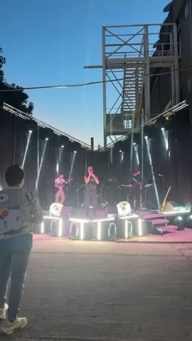 The video shows a live music performance outdoors at dusk. In the foreground, from a rear view, a person wearing a denim jacket with a colorful, possibly embroidered design and light-colored jeans stands facing the stage. Their head is turned slightly to the left. Beside them, closer to the bottom edge of the frame, a person wearing a black t-shirt with a logo and a black baseball cap with a similar logo is also facing the stage. This person's arms are down by their sides, and they are holding what appears to be a microphone or a device in their right hand.

The stage is elevated and has a modern design with bright, white LED strip lighting around its edges and base. On the stage, a band is performing. A male vocalist, wearing a black sleeveless t-shirt and dark pants, is center stage, holding a microphone and singing. To his left (stage right), a guitarist in a white shirt and khaki pants plays a guitar. Behind the vocalist, a drummer is visible behind a drum set. To the vocalist's right (stage left), another guitarist in dark clothing plays a guitar. The stage is backed by dark curtains, and the performance is illuminated by stage lights that create vertical beams of light.

Above and behind the stage, a large, industrial-looking metal structure with a staircase is visible against the twilight sky. The overall environment appears to be an open area, possibly an outdoor venue or courtyard, with a ground surface that looks like gravel or dirt. The sky is a deep blue, indicating the transition from day to night.
