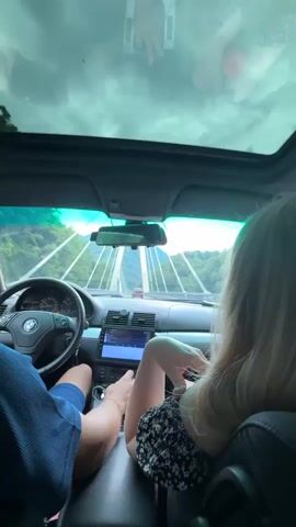 The video shows the interior of a car from the perspective of a passenger. The driver, a man wearing a blue shirt, is visible from the chest down, operating the steering wheel and gear shift. Next to him, a woman with blonde hair is seated, facing forward, with her hand near her face. The car has a sunroof that offers a view of a cloudy sky and a landscape of green hills and trees. A bridge with tall towers and cables is visible ahead, and the car appears to be driving across it. The car's dashboard and infotainment screen are also visible. Another car is seen in the distance on the bridge. The overall scene suggests a road trip through a scenic mountainous area.