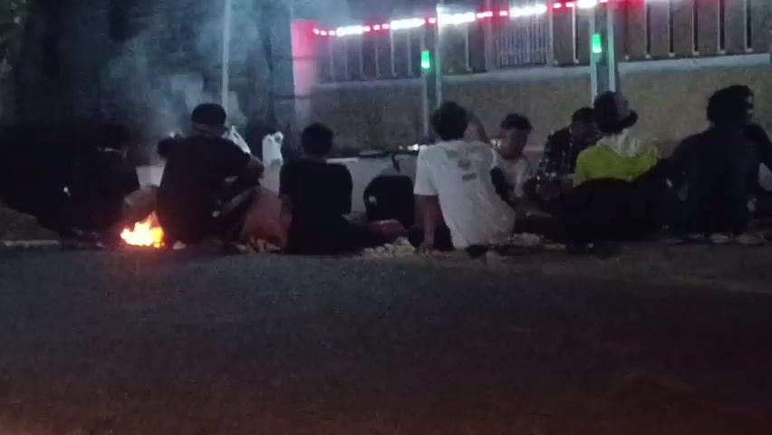 A group of young people sit around a small bonfire at night. There are at least seven individuals visible, most of them facing the fire. They appear to be sitting on the ground on a paved surface, possibly a road or sidewalk. In the background, there is a building with lights and a fence. Smoke from the bonfire drifts upwards, partially obscuring the scene. Some individuals are holding objects, though their details are unclear due to the low light and image quality. The overall atmosphere is casual and relaxed.