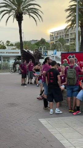 A group of people, predominantly wearing maroon t-shirts, are gathered on a paved walkway lined with palm trees. Several people are standing, while others are seated on a low wall. In the background, a building with balconies and palm trees can be seen. A sculpture of a dragon is visible on the left side of the frame. The sky appears to be sunset.