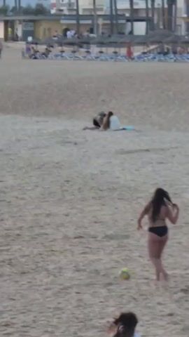 A wide shot shows a sandy beach with people scattered across it. In the foreground, a woman in a black bikini kicks a yellow and green ball. To her left, a man in blue swim trunks stands looking towards the ball. Further back, a woman in a black dress runs towards the left side of the frame, and to her left, a man in blue shorts and a white cap is holding a blue object. In the midground, two people sit on a blue mat. In the background, several beach umbrellas are set up on the sand, and buildings and palm trees are visible beyond them. Other beachgoers are visible in the distance.