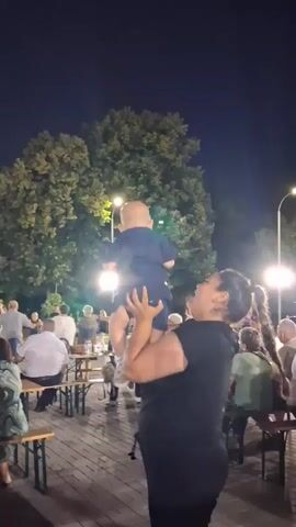 A woman with long dark hair, wearing a black t-shirt, holds a baby boy wearing a blue outfit and blue sandals in the air. The woman is smiling and looking up at the baby. The baby is facing away from the camera. In the background, there are people sitting at tables and trees with lights. The scene appears to be an outdoor gathering at night.
