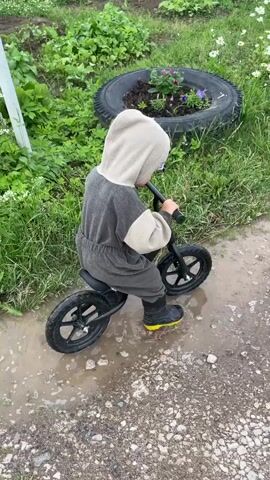 A child wearing a grey hooded jumpsuit and black boots with yellow soles rides a black balance bike through a muddy puddle. The child is facing away from the camera, and their hands are on the handlebars. The background is a grassy area with some green plants and flowers in a tire planter. The ground is wet and appears to be dirt and gravel.