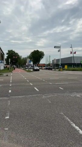 The video shows a moderately busy street intersection under a cloudy sky. In the foreground, there is asphalt road with white dashed lines marking lanes and a pedestrian crossing. On the left side of the road, a sidewalk is visible with a person walking, and further down the street, trees line the road. On the right side of the intersection, a large building, possibly a store, with a dark gray facade and green accents is visible. A few cars are driving through the intersection. The overall scene depicts a typical urban environment.