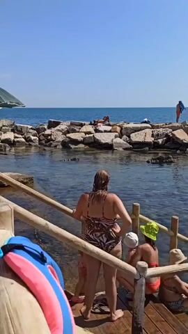 The video opens on a sunny day at a rocky beach. Several people are visible. In the foreground, a woman in a patterned swimsuit stands at the top of wooden stairs leading down to the water. Next to her, two children and another person are seated. Further down the stairs, a person in a black bikini and shorts walks into the water. In the middle ground, several people are interacting near the edge of the water, with two women in bikinis talking. Beyond them, a breakwater made of large rocks separates the calmer swimming area from the open sea. A few people are sunbathing on the rocks, and two men in swim trunks stand on the breakwater. In the background, the vast expanse of the blue sea stretches to the horizon, with a distant coastline visible. The water is clear, revealing a rocky seabed. As the video progresses, the camera pans across the water, showing more people swimming and enjoying the ocean. The scene is serene and depicts a typical summer day at the beach with various individuals engaging in water activities.