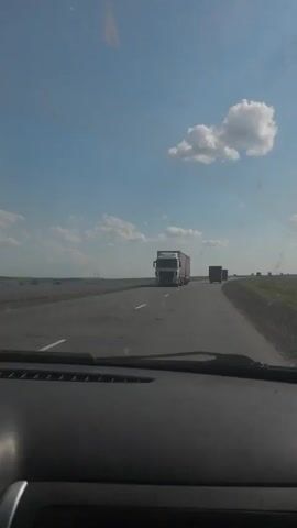 The video is filmed from inside a vehicle, looking forward through the windshield. The vehicle appears to be on a highway. There are two semi-trucks visible on the road. The first truck is in the foreground, in the left lane, and the second truck is further down the road in the right lane. The sky is blue with scattered white clouds, and the sun is shining. The landscape on either side of the road appears to be open fields. The camera then pans to the right, showing a side mirror and the passing scenery, which includes more fields and trees in the distance. The video ends with a view of the sky and the fields.