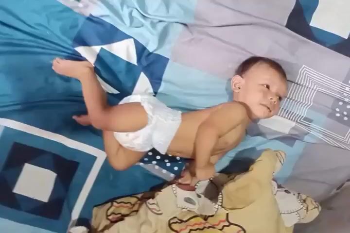 A baby wearing a diaper is lying on a bed with blue and white patterned sheets. The baby is positioned on its stomach and appears to be pushing itself up with its arms. The baby's head is turned to the side, and its eyes are open. The baby's legs are bent at the knees, and its feet are visible. There is a yellow object partially visible in the foreground, possibly a blanket or pillow.