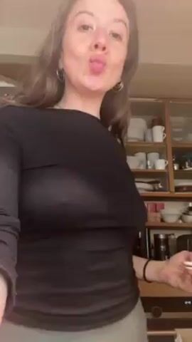 The video shows a woman in a kitchen. She's wearing a black long-sleeved shirt and light green leggings. Her hair is brown and she's wearing hoop earrings. She makes a kissy face toward the camera and then raises her arm above her head, flexing her bicep and rotating her body to the side. The background includes a refrigerator covered with magnets and a glass-front cabinet filled with dishes and kitchen items.