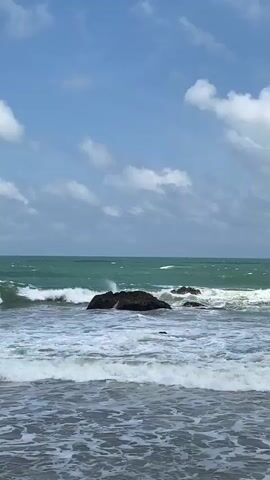The video captures a scenic view of the ocean on a cloudy day. The turquoise water is rough, with waves crashing against dark rocks protruding from the sea. The sky above is a blend of blue and white, dotted with fluffy clouds that cast soft shadows on the water. The camera focuses on the dynamic interplay between the sea and the rocks, emphasizing the power and beauty of nature. The foreground shows the foamy surf rolling towards the shore, creating a sense of movement and depth. There are no people or man-made objects visible in the frame.