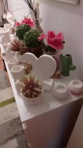 The video pans across a collection of decorative items on a white dresser. The items include various white ceramic pots and vases, some with floral or textured designs. There are also artificial plants, including succulents and a cactus, as well as faux flowers in pink and green. A white heart-shaped decorative piece is prominent in the center of the display. The lighting is dim, suggesting it might be nighttime or an indoor setting with low light. The camera movement is smooth, showing a slow sweep of the dresser's surface and the items arranged on it.