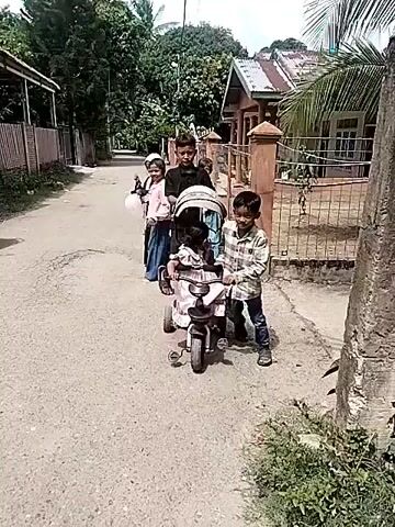 A group of children and a baby are on a street. A baby is in a pink stroller that is being pushed by a young boy wearing a plaid shirt and blue jeans. To the left of the stroller, another child wearing a pink dress and a white head covering is holding a white balloon. Behind the stroller, a boy in a black shirt is pushing the stroller. The street appears to be unpaved and has a rough surface. On the right side of the street, there is a fence and a house. The environment appears to be a rural or suburban area with vegetation and trees visible. The overall scene depicts a family outing or a group of children playing together.