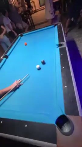 The video shows a billiards game in progress on a bright blue pool table. An orange ball, a black ball, and a white cue ball are visible on the table. A person is holding a pool cue, seemingly preparing to take a shot. People are standing around watching the game, some are sitting at a bar in the background. The setting appears to be a bar or entertainment establishment, with music playing. There's a festive atmosphere.