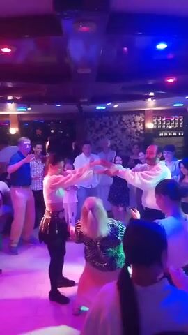 A group of people are dancing on a floor illuminated by colorful lights in a dimly lit room. Two young women are at the center of the dance floor, holding hands and dancing together. A man in a white shirt and a woman in a white top are also dancing near the center. Other people are standing and watching them dance, some of them clapping their hands. The room appears to be a restaurant or club with decorative walls and a ceiling with lights.