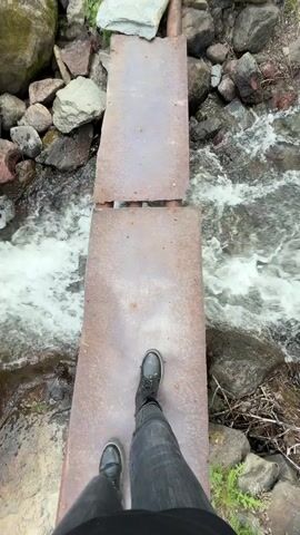 A person in jeans and black boots is standing on a makeshift bridge consisting of two rusty metal plates over a fast-flowing stream. The stream is surrounded by rocks of various sizes. The camera is positioned looking down at the person's feet, creating a first-person perspective.