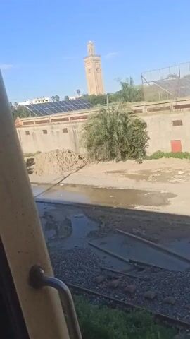 The video shows a view from inside a moving train, looking out the window at a landscape. The train tracks are visible near the bottom of the frame, lined with green vegetation. A shallow, muddy river runs parallel to the tracks.  On the other side of the river is a low concrete wall with several rectangular openings and a large pile of sand or dirt against it.  Solar panels are visible on the roof of a building behind the wall.  A tall, minaret-style tower stands prominently in the background under a clear blue sky. The camera moves slightly, affecting the angle of the view and causing a shadow to momentarily appear over the river.