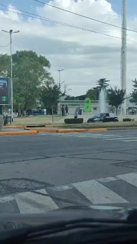 The video shows a town square in front of an industrial facility.  A fountain in the center of the square sprays water into the air.  A large, green, stylized number 2 is visible in front of the fountain.  Trees grow behind the fountain.  People are walking around the fountain.  Cars drive on the street in front of the square.  A tall, white tower is visible in the background.  The sky is cloudy.