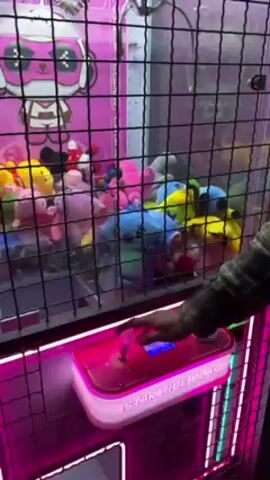 A person uses the joystick to operate a claw machine, which is lit up with pink neon lights. The claw machine is full of stuffed animals. The lower half of the machine has the words "STAR TIDE PLAY" printed on it. The machine is encased in a black wire cage.