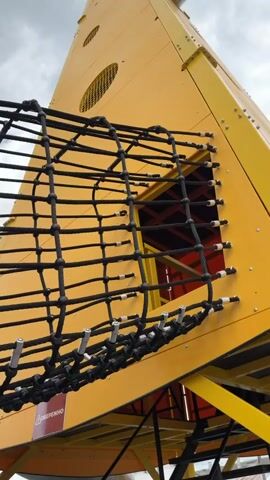 A wide shot shows a young girl in a pink vest and dark pants climbing a black rope net attached to a large yellow structure. She is moving from left to right across the frame. The yellow structure appears to be a playground element, possibly a climbing tower or slide, with an opening visible. The sky is cloudy. The camera is positioned below the girl, looking up at her and the playground. The video appears to be shot outdoors.
