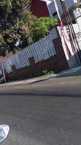 A first-person perspective video shows a ride on a vehicle, likely a motorcycle, down a paved road on a sunny day. The camera is angled slightly downwards, capturing the road and the right side of the environment. Along the right side of the road, there is a sidewalk and a low brick wall topped with a white picket fence. Beyond the fence, several trees with green foliage are visible. Further down the road, there are buildings with brown brick bases and white walls, and some have awnings. Some of the buildings appear to be small shops or stalls, with one displaying a sign that says "BACOK" and another that lists various food items. There are also poles with electrical wires running overhead, and several Indonesian flags are visible, attached to poles and flying in the breeze. The road has white lane markings. The vehicle appears to be moving at a moderate speed. The overall impression is that of a ride through a residential or commercial area in a warm climate.