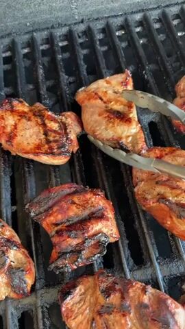 The media file shows a close-up, overhead view of several pieces of seasoned meat being grilled on a black metal grill. The meat pieces are various shapes and sizes, with visible grill marks and browning indicating they are cooking. A pair of silver metal tongs is used to turn one of the pieces of meat. The grill grates are black and appear to be well-used. The background is out of focus but suggests an outdoor setting. There is no visible text. The overall impression is of food being prepared for a meal.