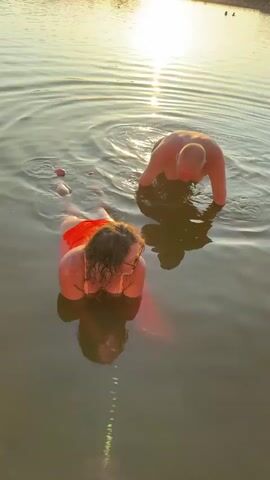 The video shows two people in shallow water at sunset. A woman is lying on her stomach in the foreground, wearing an orange garment and sunglasses. Her legs are visible above the water, and a red object, possibly a buoy, is attached to her ankles. Behind her, a man is also in the water, with his upper body visible and his head tilted down as if he is speaking or looking at her. The water is calm with ripples, and the sun is reflected on the surface. The overall scene is of a relaxed, recreational moment in a natural body of water.