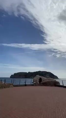 The video shows a coastal scene with a rocky island in the distance under a blue sky with wispy clouds. The camera pans to the left, revealing a red brick paved area with a metal railing along the edge. A large rock formation is visible on the side. The camera continues to pan, revealing three people sitting on a bench overlooking the sea. The people are wearing warm clothing.