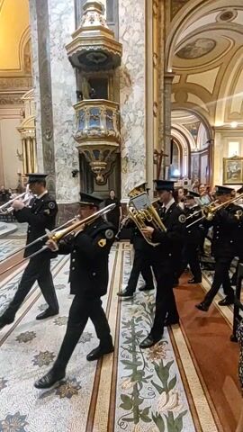 A police band plays in a church. The band is marching down the aisle toward the camera while playing. The band members are dressed in dark-blue uniforms with dark hats. They are playing instruments like a tuba, saxophone, trombones, and a flute. Spectators are standing on either side of the aisle watching the band march. Some are recording the band with their phones. The church has marble walls, and arched ceilings with ornate details. The floor is also marble and has an elaborate floral pattern down the aisle. There is a golden pulpit attached to one of the marble columns.