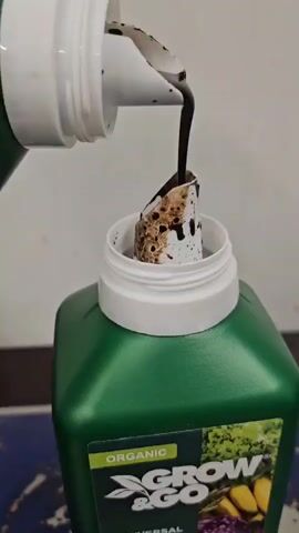 A close-up shot shows dark liquid being poured from a white bottle into a green bottle. The liquid is thick and viscous, with some small dark particles suspended in it. The green bottle has a label that says "ORGANIC GROW & GO". The liquid is pouring into the green bottle's opening, which has a white cap with a cone-shaped insert. Some of the liquid has spilled and dripped onto the insert and the inside of the green bottle's opening. The background is a plain, light-colored wall.