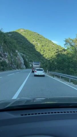 The video shows a view from inside a car driving on a paved road. The car is following a white hatchback, which is in turn following a bus. The road is surrounded by lush green mountains. The sky is clear and blue. The sun is casting shadows on the mountainside, indicating it is likely morning or late afternoon. The road has white lane markings and a guardrail on the right side. The white hatchback has a license plate visible. The bus appears to be a passenger bus. The video is shot from a first-person perspective, as if the viewer is the driver of the car.