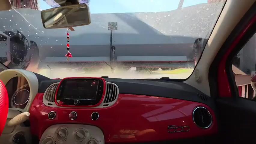 The video provides an interior view from the driver's seat of a red Fiat 500, looking out the windshield. The car is inside an automatic car wash. Water is spraying from overhead equipment, creating a mist that obscures the view outside. On the right side of the frame, red brushes can be seen as part of the car wash machinery. The dashboard of the car, with its red accents and central infotainment screen, is clearly visible. A rear-view mirror hangs from the windshield with a beaded charm attached. The car is moving forward through the car wash.