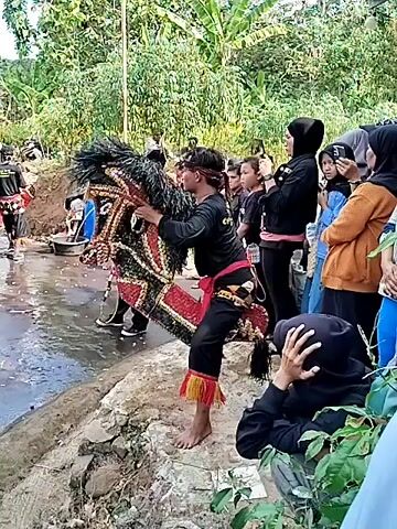 A group of people are gathered outdoors, some of whom are observing an outdoor cultural performance. A young person in traditional attire, wearing a mask of a horse's head, is the central focus of the performance. They are dressed in black clothing with a red sash and have a decorated headdress. Behind the performer, other individuals are visible, some wearing traditional clothing, and one person is playing an instrument. The environment appears to be a village setting with lush green vegetation and trees in the background. The ground is uneven and wet, suggesting it may have recently rained or is near a water source. The people are positioned around the performer, watching and some are capturing the event on their phones. The overall atmosphere seems festive and communal.