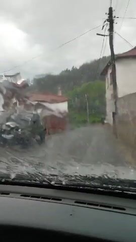 The video shows a view from inside a vehicle, looking through the windshield. It appears to be raining heavily, as water is streaming down the windshield, obscuring the view of the outside. The windshield wipers are active, clearing some of the water. The scene outside the vehicle depicts a street in a village or rural area. There are buildings on either side of the street, some with red tile roofs. Trees and a hillside are visible in the background. The street itself appears to be flooded or covered in a significant amount of water and debris, suggesting recent heavy rainfall or a possible mudslide. The overall atmosphere is gloomy due to the overcast sky and the heavy rain. There are no people clearly visible in the frame, nor any other vehicles actively moving. The focus is on the challenging driving conditions due to the weather.