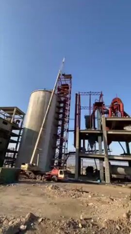 The video shows an industrial site with several large structures and machinery under a clear blue sky. A crane is positioned next to a tall cylindrical silo, which has a metal structure with stairs attached to its side. Next to the silo is a multi-level structure with red and gray components, seemingly part of the industrial complex. The ground appears to be dirt and gravel. There is a shipping container on the ground and some other construction materials. The crane's boom is extended towards the top of the metal structure attached to the silo. No people are clearly visible in the shot.