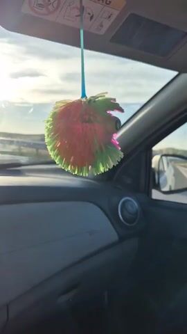 The video shows a fuzzy, multicolored ball hanging from the visor inside a car. The ball is predominantly pink and green. Sunlight streams in from outside, casting shadows and highlights on the car's interior. The dashboard, air vent, and side mirror are visible. Outside the car, a road and a light-colored sky can be seen, suggesting the car is in motion on a sunny day. A safety warning sticker with pictograms, including an airbag symbol, is visible on the visor.