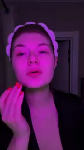 A woman in a black robe and a white headband is applying a white cream to her face in a room lit with pink light. Her fingernails are painted red. She is looking directly at the camera. The background includes a white curtain, a door, and a piece of furniture. She uses her right hand to rub the cream into her skin.