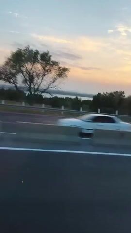The video shows a highway at sunset. The sky is a mix of orange, yellow, and blue with scattered clouds. There are trees and a body of water in the distance. A concrete barrier divides the highway lanes. A pedestrian bridge is visible over the highway. The camera is moving, giving a sense of travel. The overall impression is of a scenic drive at dusk.