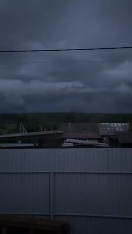 The video shows a dark, cloudy sky over a rural landscape. In the foreground, there is a light gray metal fence. Behind the fence, there are several buildings with corrugated metal roofs, some appearing weathered. Trees form a dark line on the horizon, and the sky is filled with heavy, dark clouds, suggesting an approaching storm. The lighting is dim, and the overall atmosphere is somber and overcast. There are no people or animals visible in the video.