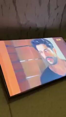 The video shows a television screen playing a scene from a movie or TV show. The scene features a man and a woman, the woman wearing a blue shirt and the man in a black t-shirt. They appear to be in a conversation. The television is placed on a surface, likely a bed or a sofa. The background wall has a marble-like pattern. The ZOR logo is visible on the top right corner of the screen.