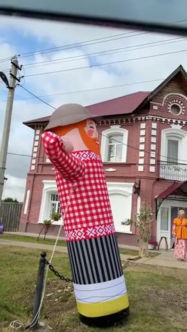 An inflatable dancing tube man stands on a grassy lawn in front of a two-story red brick house with white trim. The house has a red roof and several windows. Three women are standing on the porch of the house. The woman on the left is wearing a bright orange and pink patterned outfit and a headscarf. The woman in the middle is wearing a green and yellow striped dress with a decorative necklace and a headscarf. The woman on the right is wearing a pink and purple plaid dress and a headscarf. The sky is cloudy.