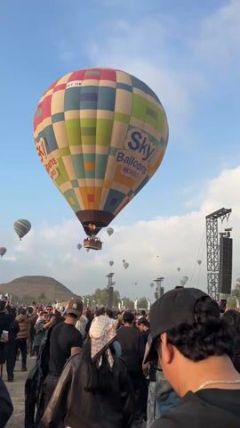 The video shows a crowd of people gathered below several hot air balloons ascending in the sky. The main balloon in the foreground is large and multicolored, with "Sky Balloons MEXICO" written on its side. People in the crowd are looking up, many holding up their phones to capture the scene. A pyramid-like structure can be seen in the background. The sky is partly cloudy.
