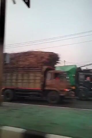 The video captures a view from a moving vehicle, possibly a car, traveling along a road. The camera is angled slightly downwards and to the right. The footage begins with a blurry shot of a truck loaded with what appears to be sugarcane or logs, its red chassis and sides visible. The truck is moving in the same direction as the camera. Next, a large white tanker truck with red accents and visible branding passes by. This is followed by a view of a distinctive red arch bridge in the distance, with mountains behind it and power lines in the foreground. Finally, the camera shows a light green box truck passing by. The overall environment appears to be urban or suburban, with signs of infrastructure like roads, bridges, and power lines. The lighting suggests it might be late afternoon or early morning. The video is characterized by motion blur due to the movement of the camera.