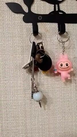 A wall-mounted black metal key rack is shown in the video. Several items hang from it, including keys, three pink plastic figures with keychains, and a gray and white stuffed cat with a keychain. The figures are cartoonish in design. The rack is attached to a wall with a textured beige covering. The camera pans slightly to the right, revealing more of the rack's contents.