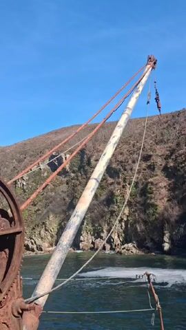 The video shows a close-up of an old, rusty crane mechanism against a bright blue sky and a hilly, rocky coastline. The crane features a large, toothed gear wheel and a boom supported by wires. The boom extends over a body of water, with white foam visible on the surface. The crane appears weathered and inactive.