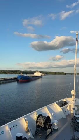 The video opens with a low-angle shot of the deck of a ship, showing a coiled rope on a spool and a white flagpole against a blue sky with scattered clouds. The camera then pans slightly to the left, revealing a large blue and red cargo ship docked at a pier in the distance. The cargo ship is long and low, with a white superstructure. The water is calm and reflects the blue sky. The background is a tree-lined shore with a few buildings visible. The scene is illuminated by the warm light of what appears to be either sunrise or sunset. The video is very still with no apparent movement other than very subtle camera drift.