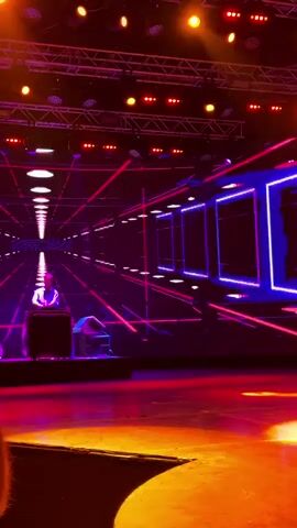 The video shows a stage with a DJ set up in front of a large LED screen. The screen displays a colorful, abstract graphic that resembles a tunnel or a futuristic cityscape. The DJ is wearing a white shirt and a hat. The stage is lit with purple and red lights. The video is shaky and appears to be filmed from a distance.