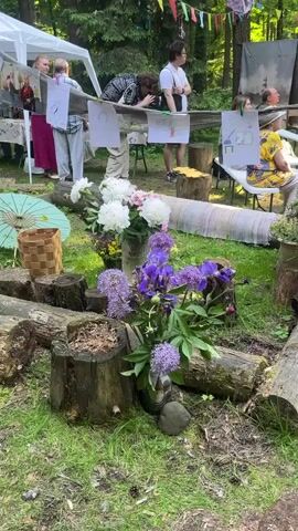The video depicts an outdoor gathering in a wooded area. In the foreground, logs are arranged in a semi-circle, with flowers, including peonies and purple alliums, placed in vases and buckets. A woven basket and a green umbrella are also visible. In the background, several people are seated on chairs, facing towards a central area. Some are observing something, while others are engaged in conversation. There are also drawings or posters displayed on a wooden structure. A white tent is set up on the left side of the frame. A Russian flag is visible, attached to a pole. The overall atmosphere suggests a community event or an informal celebration held in nature.