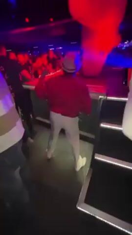 Here's a concise description of the video:

The video shows a person dancing on a platform at a nightclub. The individual is wearing a red jacket, light-colored pants, a hat, and white sneakers.  They are facing away from the camera.  Behind them, there's a large inflatable structure, illuminated with red light, moving in time with the music.  Other people are visible in the background, suggesting a crowded atmosphere.  The lighting in the club is dark, with flashing blue and red lights.